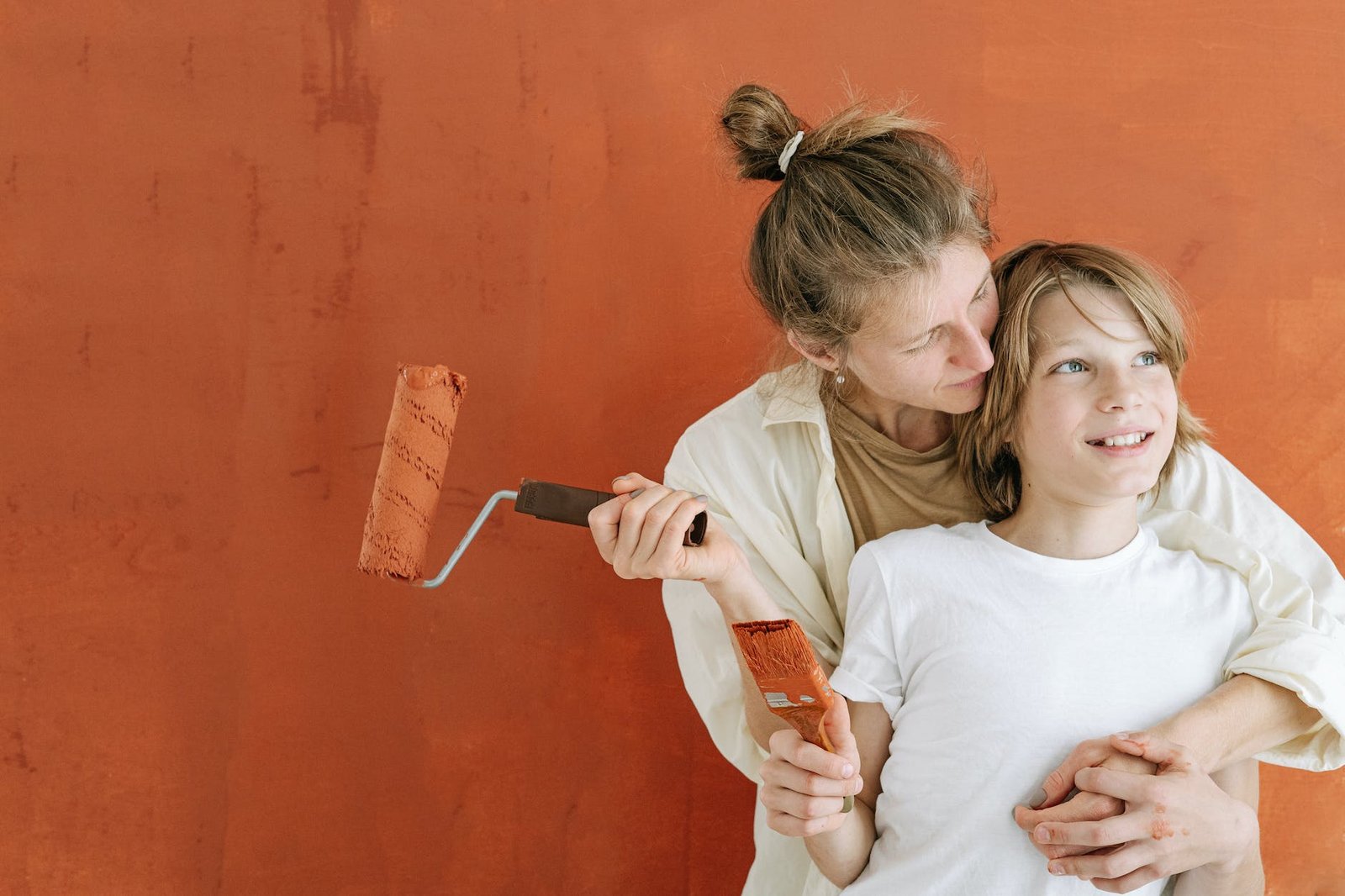 woman in white button up shirt holding a paint roller hugging a boy holding a paintbrush